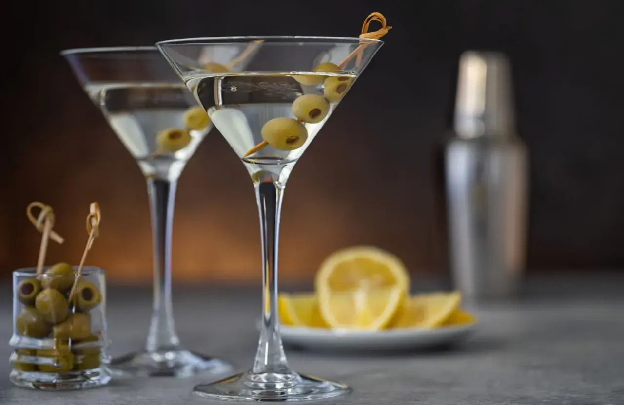 Deux verres à martini remplis, garni d'olives. Une recette de dirty martini à portée de main, avec des citrons et un shaker en arrière-plan.