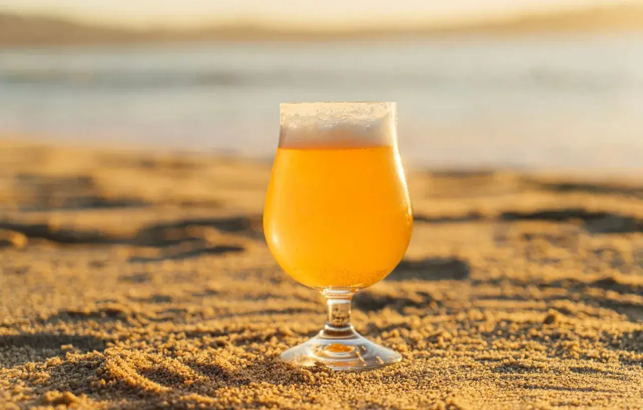 Un verre de bière blonde, type IPA, sur le sable chaud, avec la mer en arrière-plan au coucher du soleil.