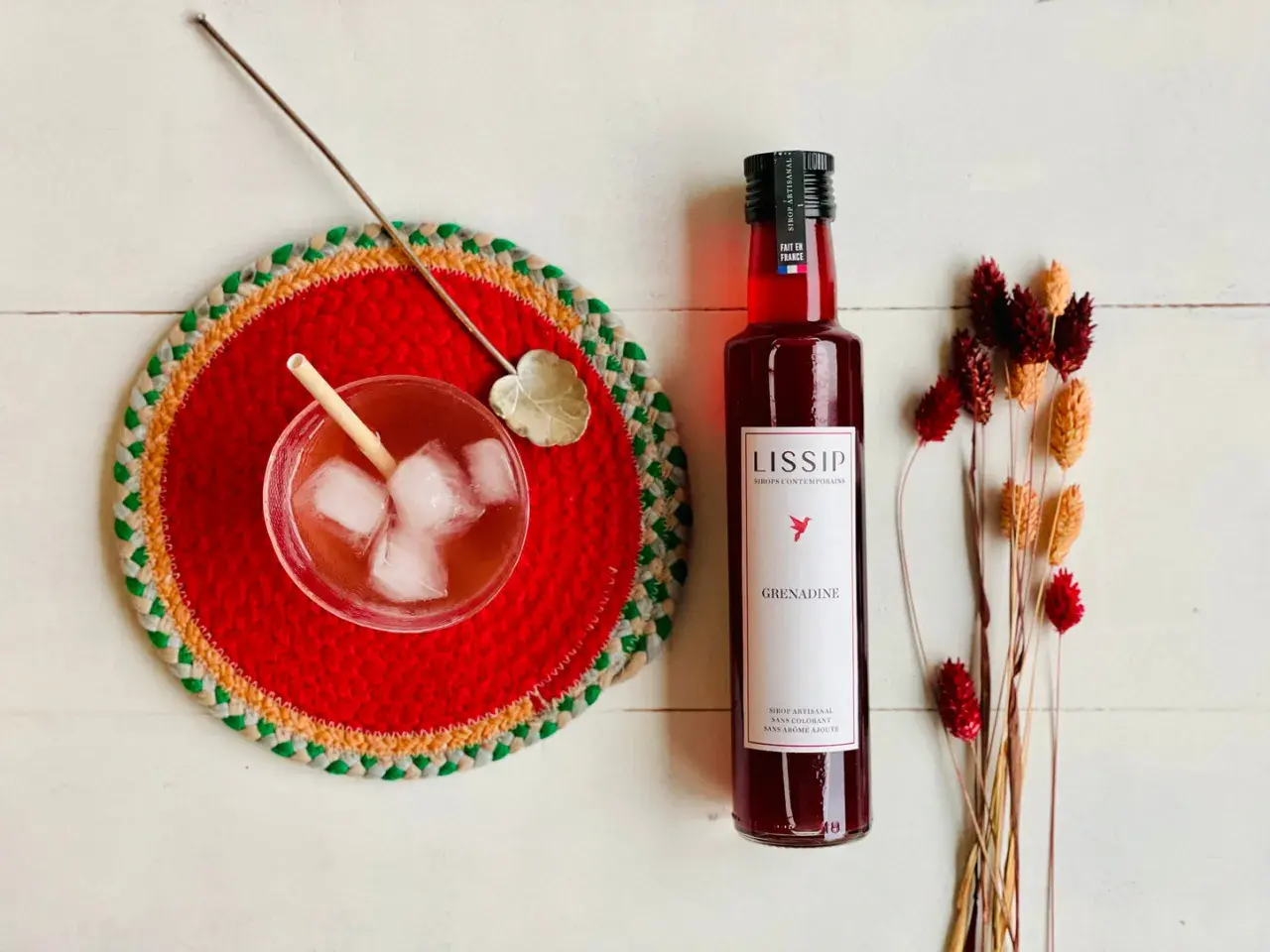 Un verre de boisson pétillante avec de la bière et de la grenadine, une bouteille de sirop de grenadine Lissip et des fleurs séchées.