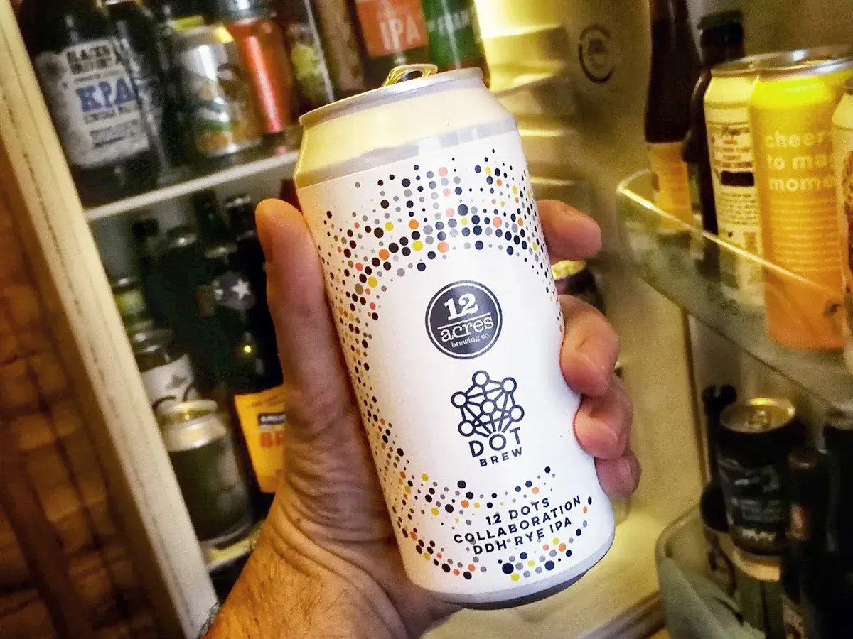 Une main tient une bière canette forte "12 Acres Dot Brew" devant un réfrigérateur rempli de bières.