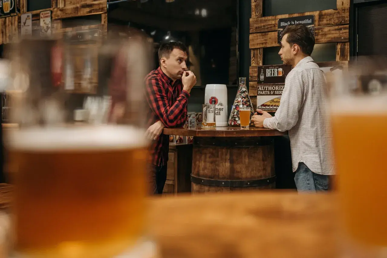 Deux hommes discutent près d'un bar en bois, une bière la bête à la main, dans une ambiance rustique.