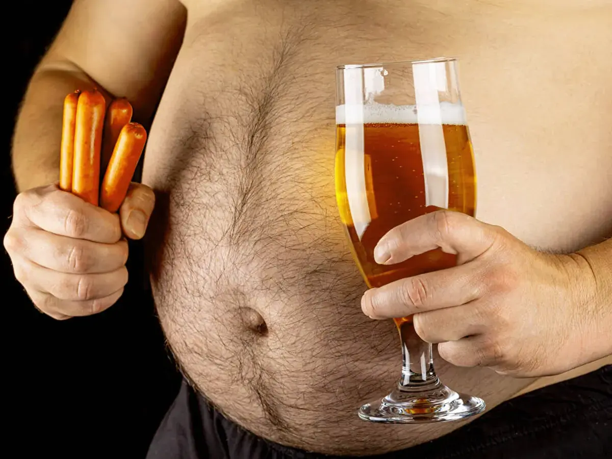 Un homme tient un verre de bière et des saucisses, soulevant la question : la bière fait elle grossir ? Son ventre proéminent suggère une réponse.