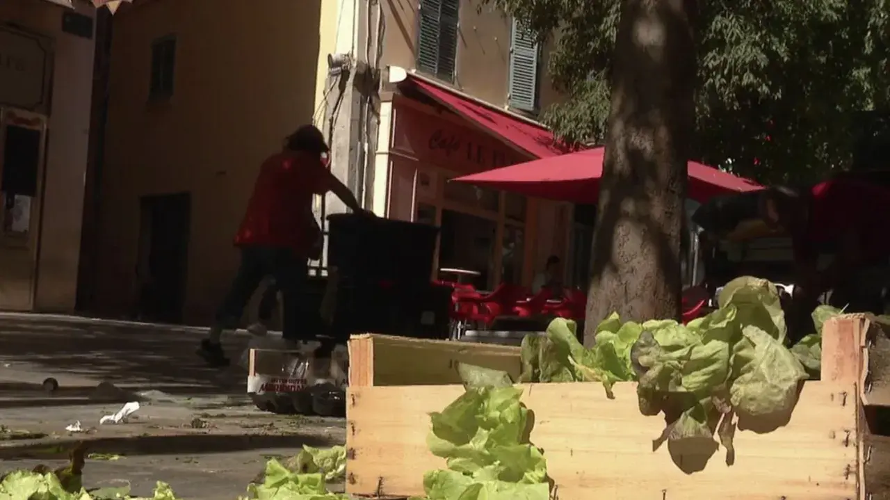Des caisses de laitues fraîches attendent sur un trottoir, tandis que des personnes chargent des bacs. La duree fut de biere, mais le travail continue.