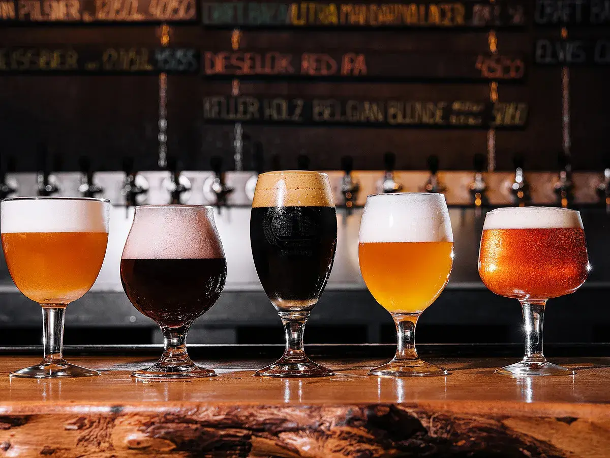 Cinq verres de bière, dont une pale ale ou ipa, sur un comptoir en bois. Derrière, des tireuses et un tableau des bières.