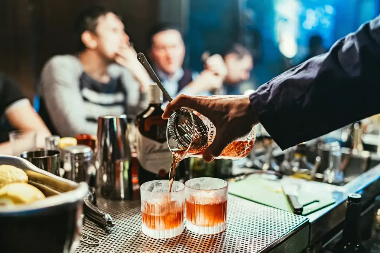 Un barman pr&eacute;pare un cocktail classique, versant un liquide ambr&eacute; dans deux verres remplis de glace. Des clients flous sont en arri&egrave;re-plan.