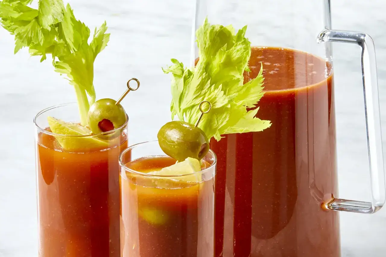 Deux verres de Bloody Mary, garni de céleri et d'olives, à côté d'une carafe remplie de la même boisson rouge vif.