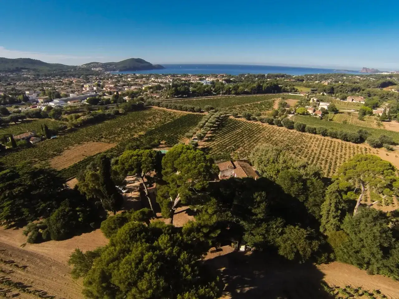 Vue aérienne du Château Pradeaux et ses vignobles s'étendant vers la mer Méditerranée, sous un ciel bleu éclatant.