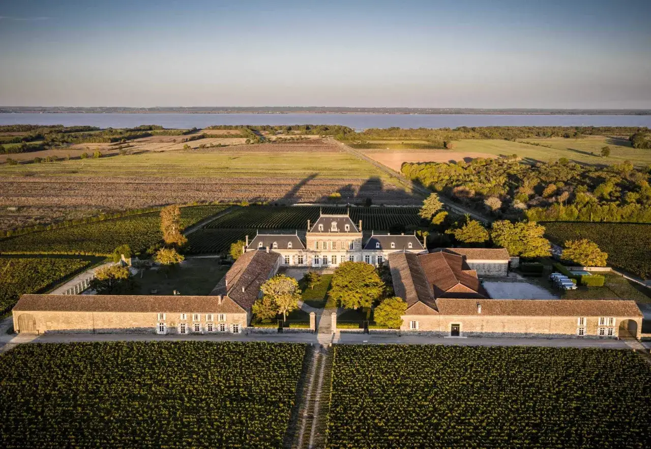 Vue a&eacute;rienne du Ch&acirc;teau Pey La Tour baign&eacute; par le soleil couchant, entour&eacute; de vignobles &agrave; perte de vue et d'une &eacute;tendue d'eau.