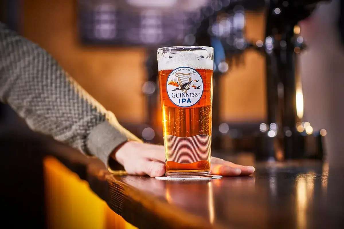 Un verre de bière IPA Guinness, ambrée et pétillante, posé sur un comptoir en bois. Une main repose à côté.