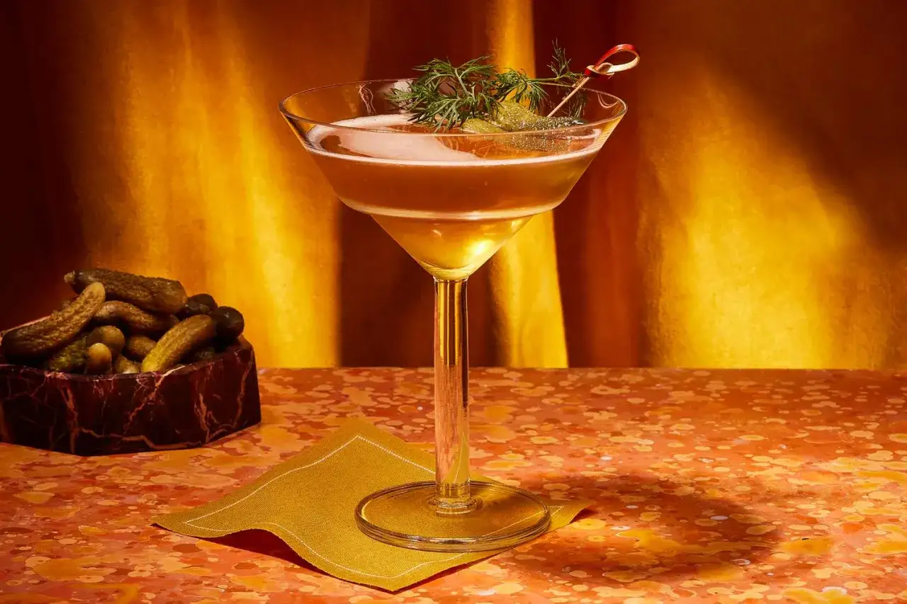 Un cocktail ambr&eacute;, garni d'aneth et de cornichons, &eacute;voque un nom de cocktail f&eacute;minin. &Agrave; c&ocirc;t&eacute;, un bol de cornichons et une serviette dor&eacute;e.