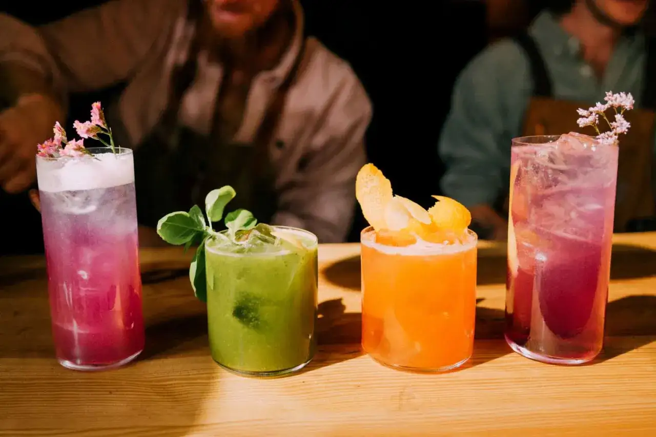 Quatre cocktails color&eacute;s, dont un cocktail 5 alcool, sont pr&eacute;sent&eacute;s sur un bar en bois. L'un est rose avec des fleurs, un autre vert avec des feuilles, un troisi&egrave;me orange avec des zestes, et le dernier violet.