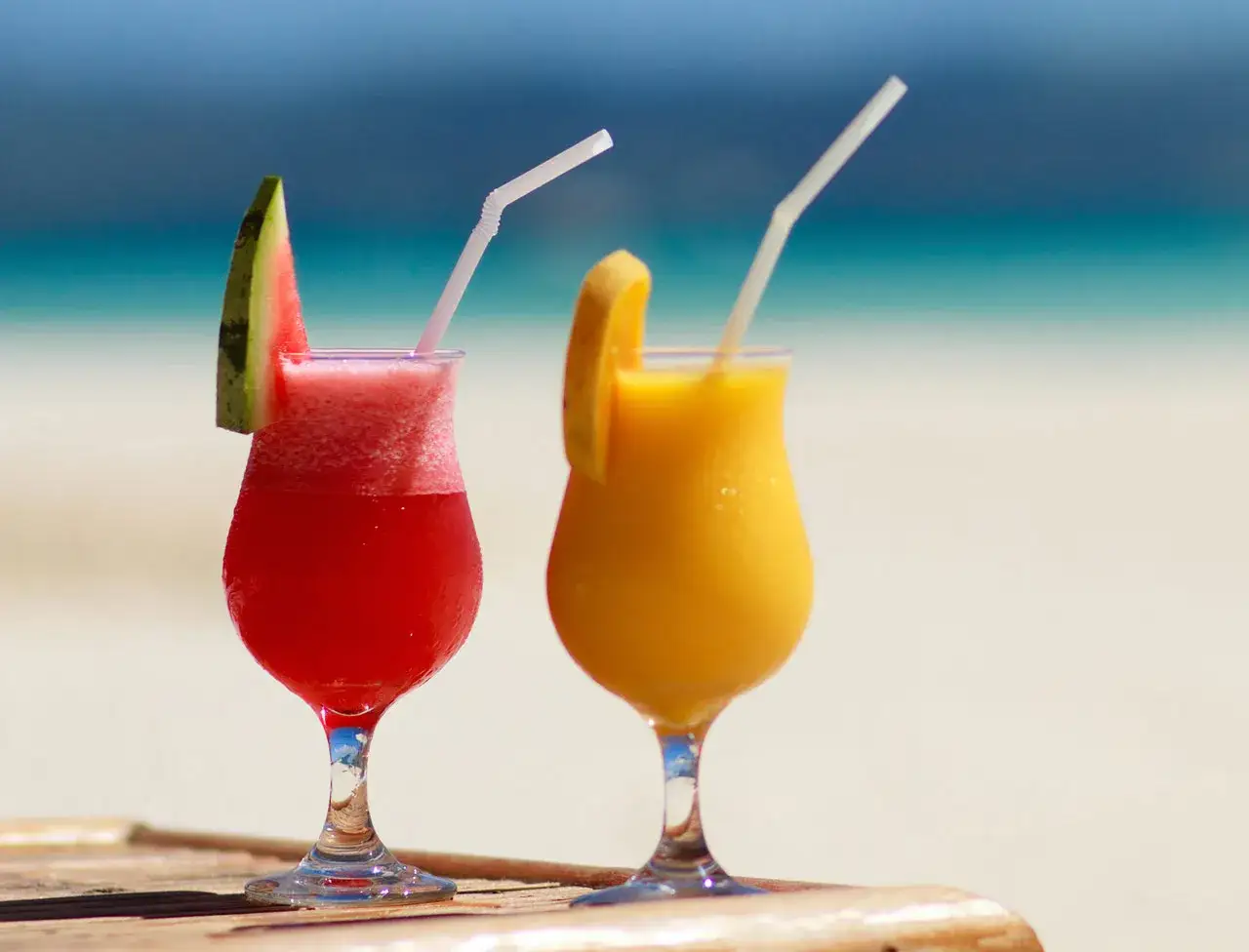 Deux cocktails d'été colorés, un rouge à la pastèque et un jaune à l'orange, sur fond de plage et de mer.