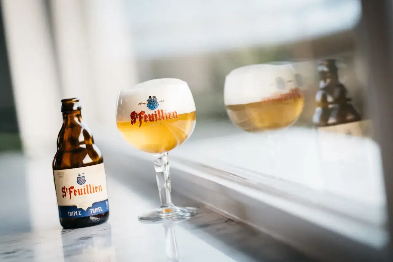 Une bouteille et un verre de bi&egrave;re triple St. Feuillien, avec une mousse g&eacute;n&eacute;reuse, refl&eacute;t&eacute;s sur une surface.