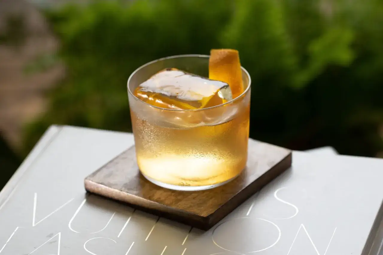 Un verre de cocktail ambr&eacute;, orn&eacute; d'un zeste d'orange et d'un gros gla&ccedil;on transparent, repose sur un sous-verre en bois.