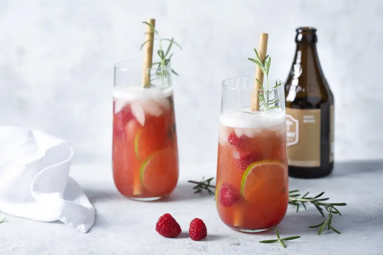 Deux verres de cocktails fruit&eacute;s, garnies de framboises et de rondelles de citron vert, avec une touche de romarin. Une bouteille de bi&egrave;re est visible en arri&egrave;re-plan.