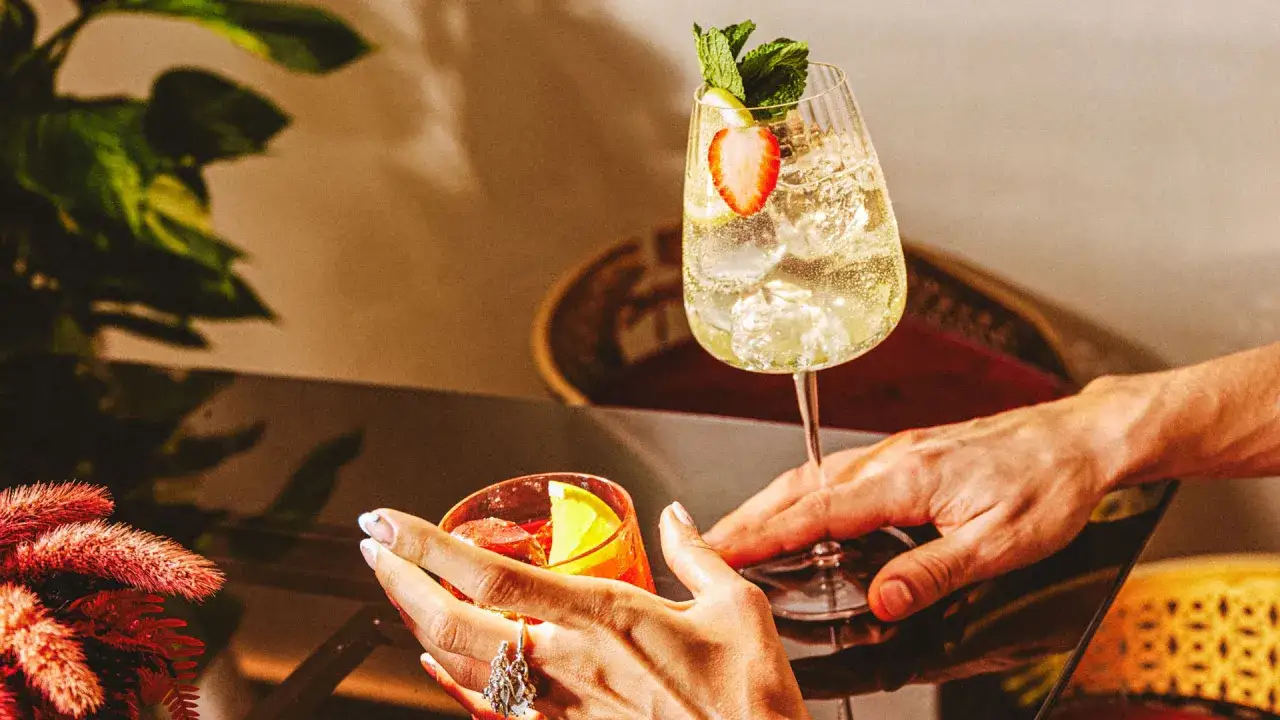 Deux mains tendent des cocktails rafra&icirc;chissants, un verre de martini bitter rouge et un autre p&eacute;tillant garni de fraises et de menthe.