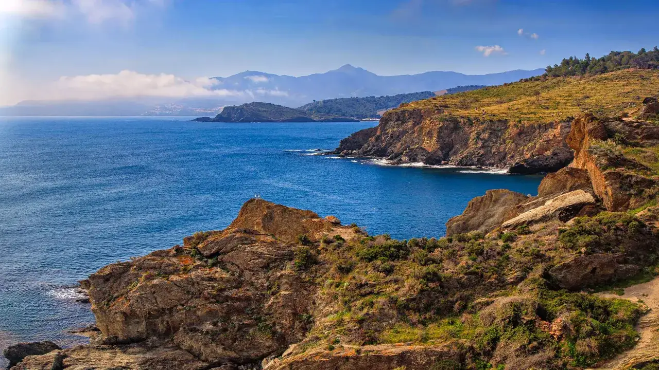 Vue panoramique des falaises rocheuses et de la mer bleue, avec des montagnes brumeuses à l'horizon. Les clos de Paulilles, un paysage côtier sauvage.